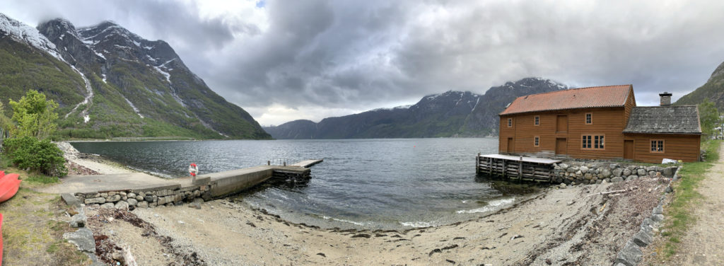 A panorama of a nordic fjord a sandy beach in the fore ground, a two storey wooden boathouse to the right. Clouds shroud mountains across the water.