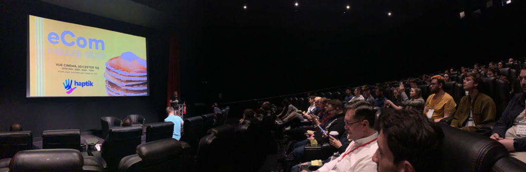Interior panorama of a full cinema - a man stands by the screen presenting a slide that reads eCom collab club