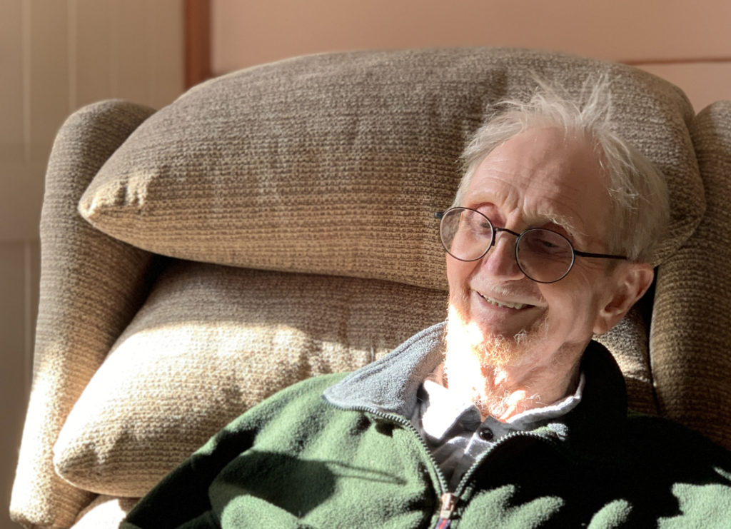 An elderly man in glasses leans back in a beige armchair, the sunlight lighting up his smiling face.