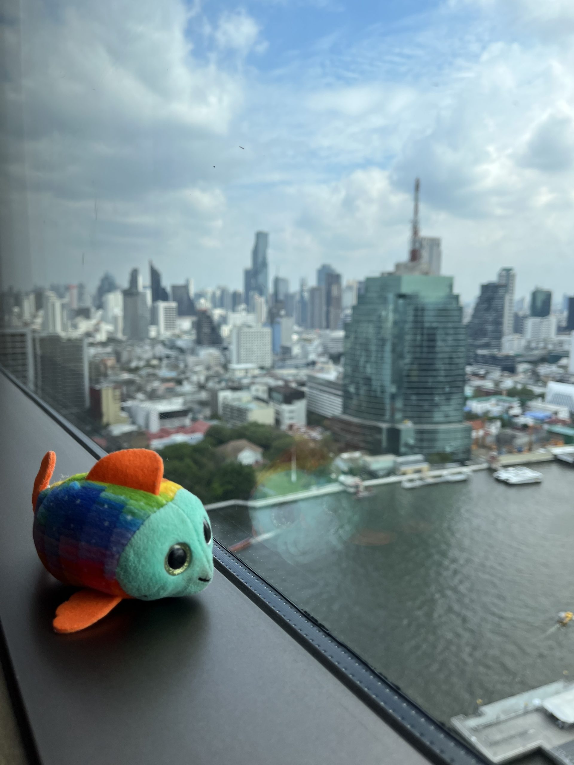 A plushy fish on a windowsill looking out over downtown bangkok
